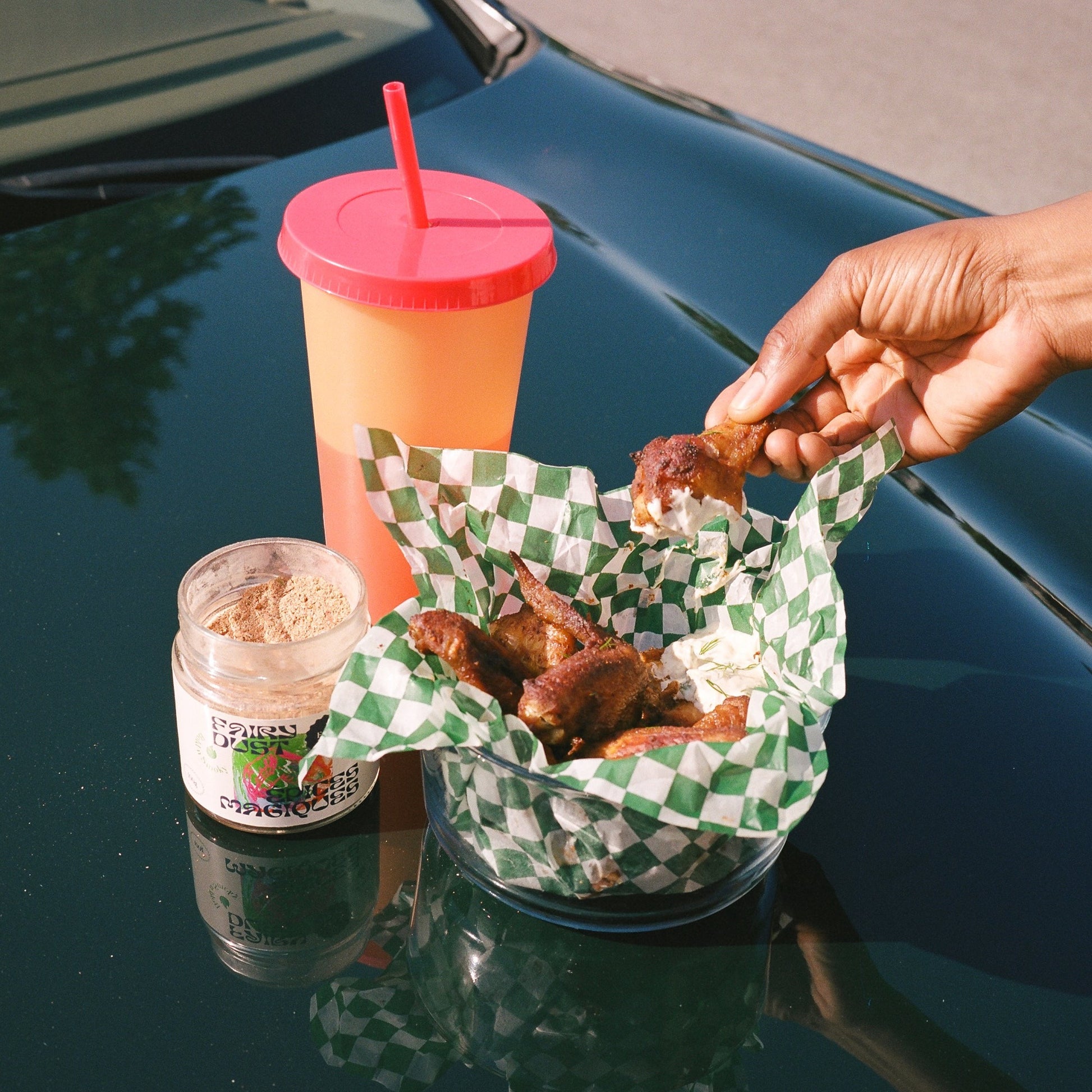 Hand grabbing for BBQ chicken wings made with ranch dip  next to on open jar of Bold n Smoky Fairy Dust spice blend and a drink on the hood of a car in summer